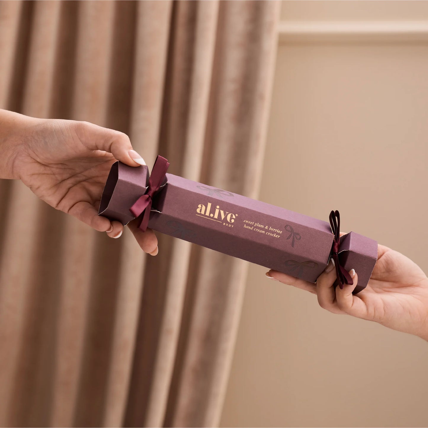 Two hands holding a purple 'al.ive' product against a beige curtain background