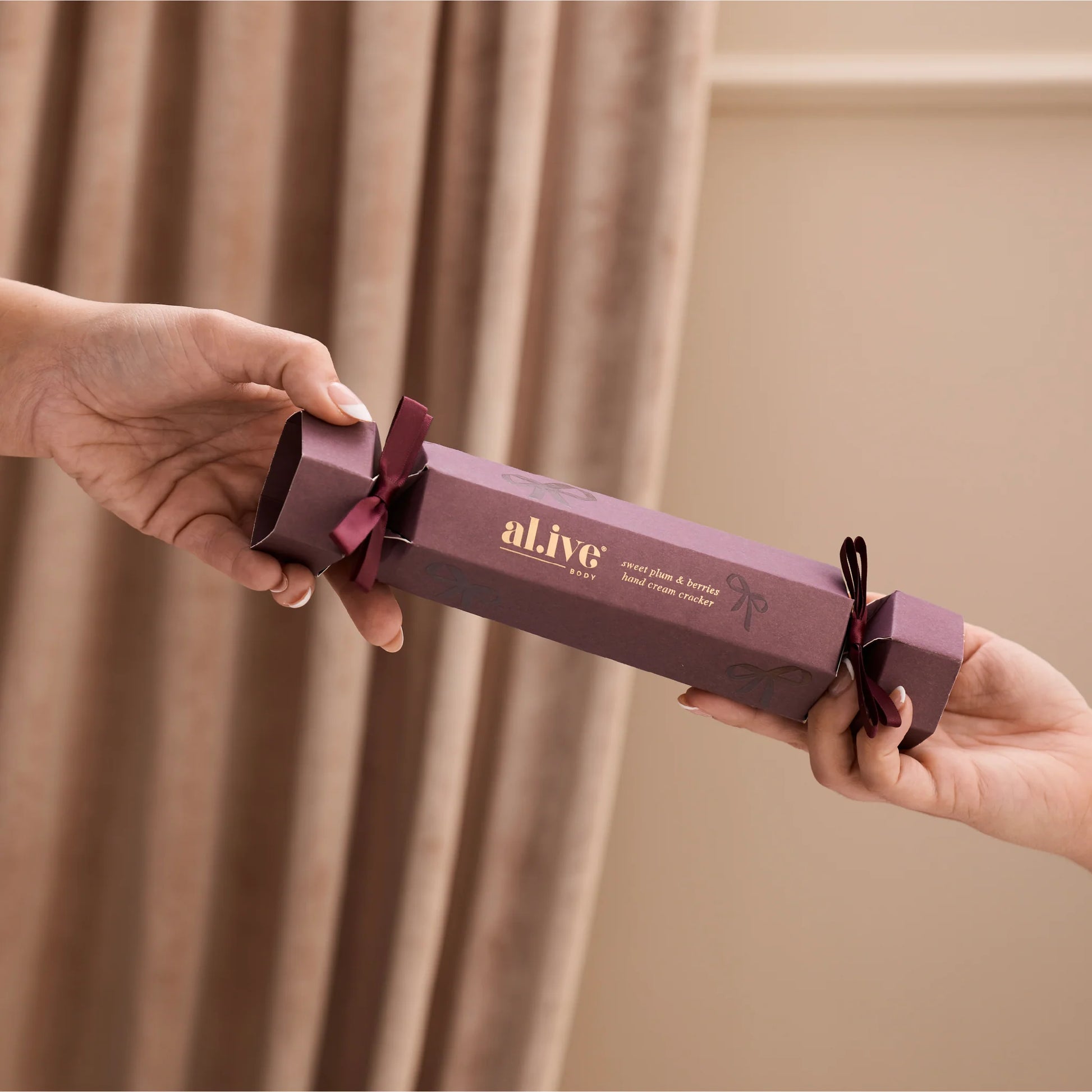 Two hands holding a purple 'al.ive' product against a beige curtain background