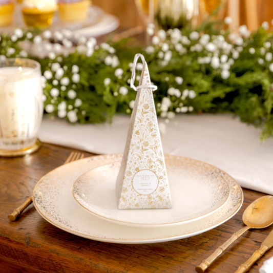 A triangular favor bag with a gold floral pattern and white hanging loop, standing on a gold-rimmed plate on a wooden table with a blurred event background.