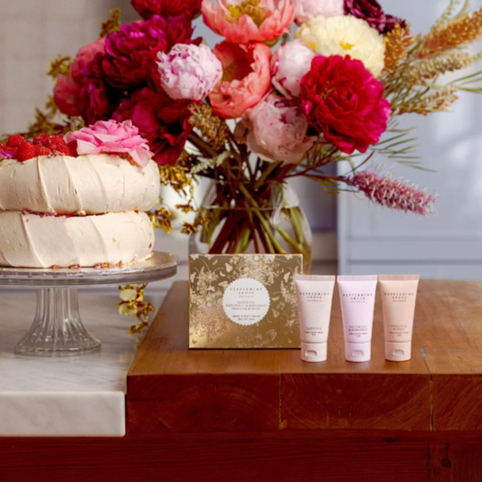 A beauty gift set featuring three pale pink tubes of Bath Body Works body wash, a gold floral box, and a small gold card, arranged on a wooden table next to a decorated cake and a large pink and red flower arrangement.