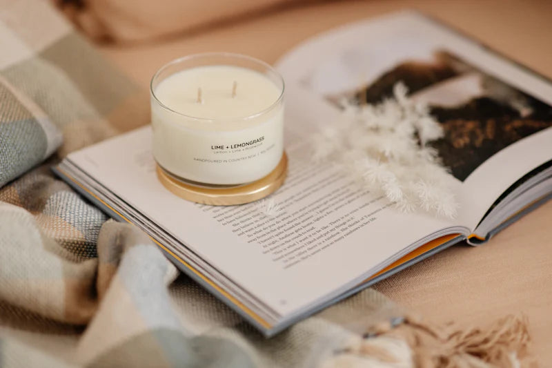 Candle with 'Lime + Lemongrass' scent on an open book, surrounded by cozy blankets and decor.
