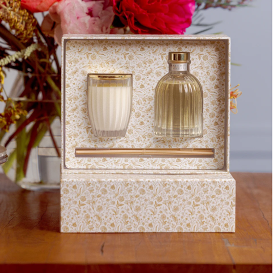 A gift set in a floral-patterned box containing a white candle in a ribbed glass holder and a clear glass bottle with a gold cap, displayed on a wooden table with flowers in the background.