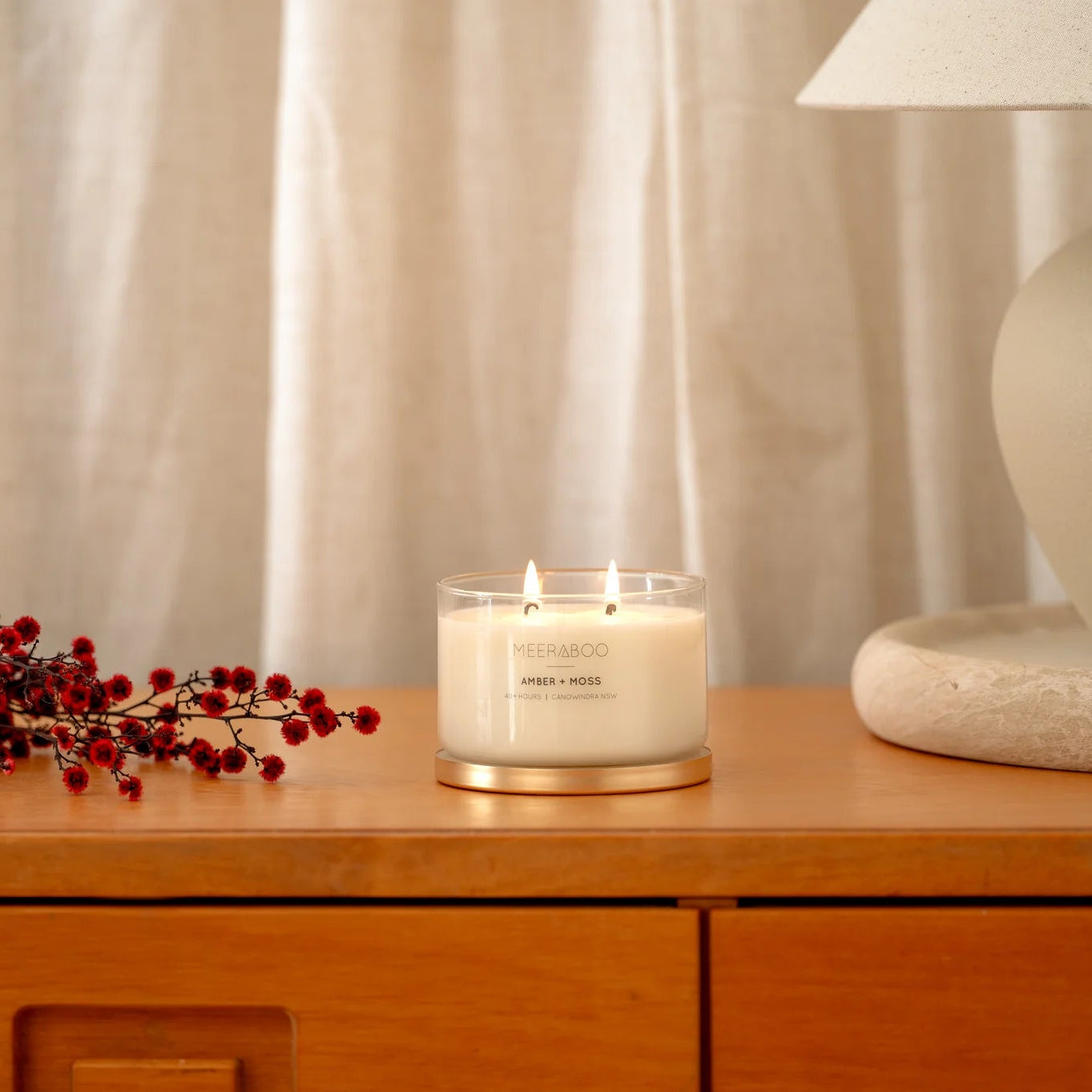 Candle on a wooden surface with a lamp and decorative branches in the background
