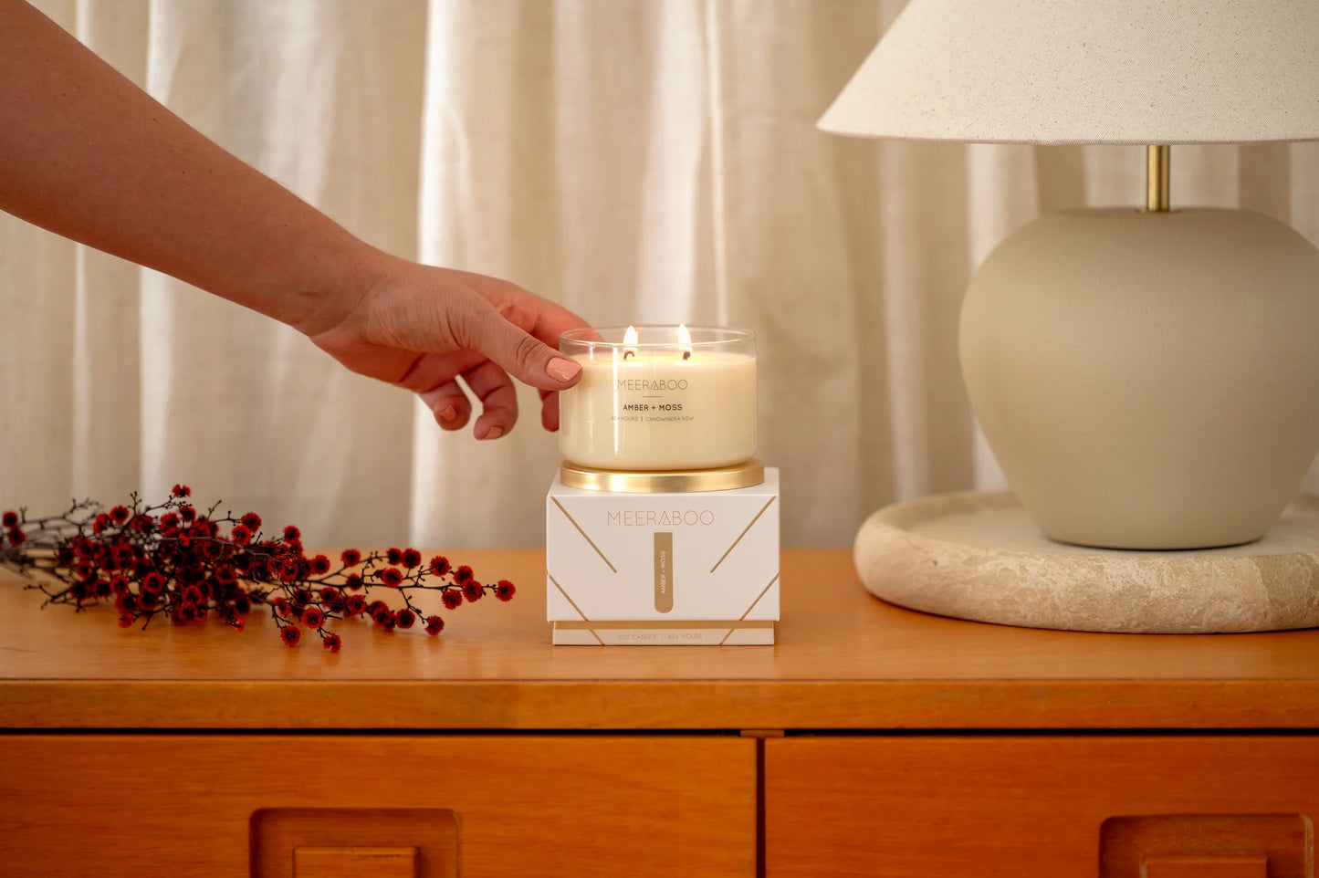 Hand reaching for a candle on a wooden surface with a lamp and decorative items in the background.