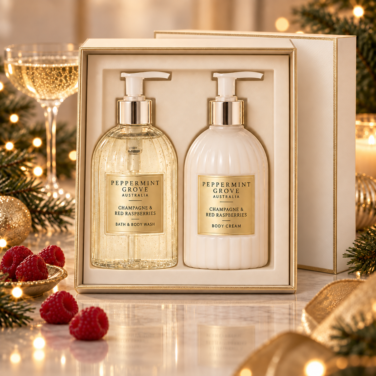 A Peppermint Grove Australia bath and body gift set in an open cream-colored box, featuring a clear bottle of champagne and red raspberry bath & body wash and a white bottle of champagne and red raspberry body cream, both with gold pump collars and gold labels. The set is styled on a reflective marble surface with festive holiday decorations, including pine branches, gold ribbons, a glass of sparkling wine, and fresh raspberries.