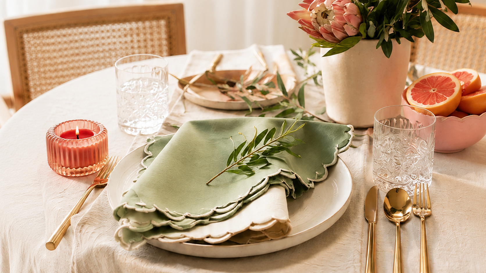 A beautifully styled round table set for a dinner party, featuring a stack of sage green scalloped napkins on white plates, gold flatware, a pink candle, and a vase of protea flowers.