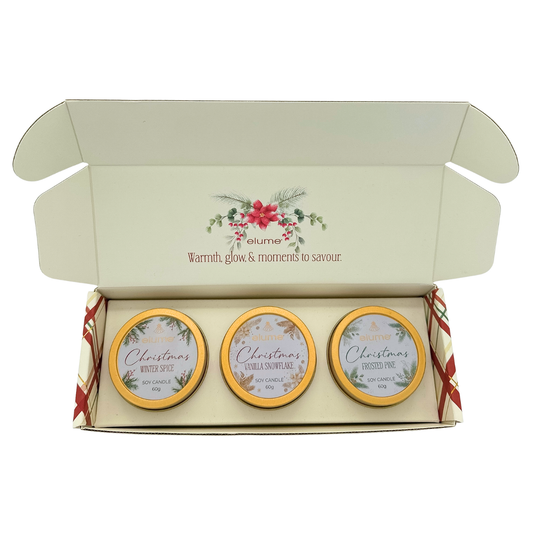 An open cream-colored gift box containing three small round candles with gold-rimmed lids. The inside lid of the box features a red and green holly illustration and the text 'volume Warmth, glow & moments to savour'.