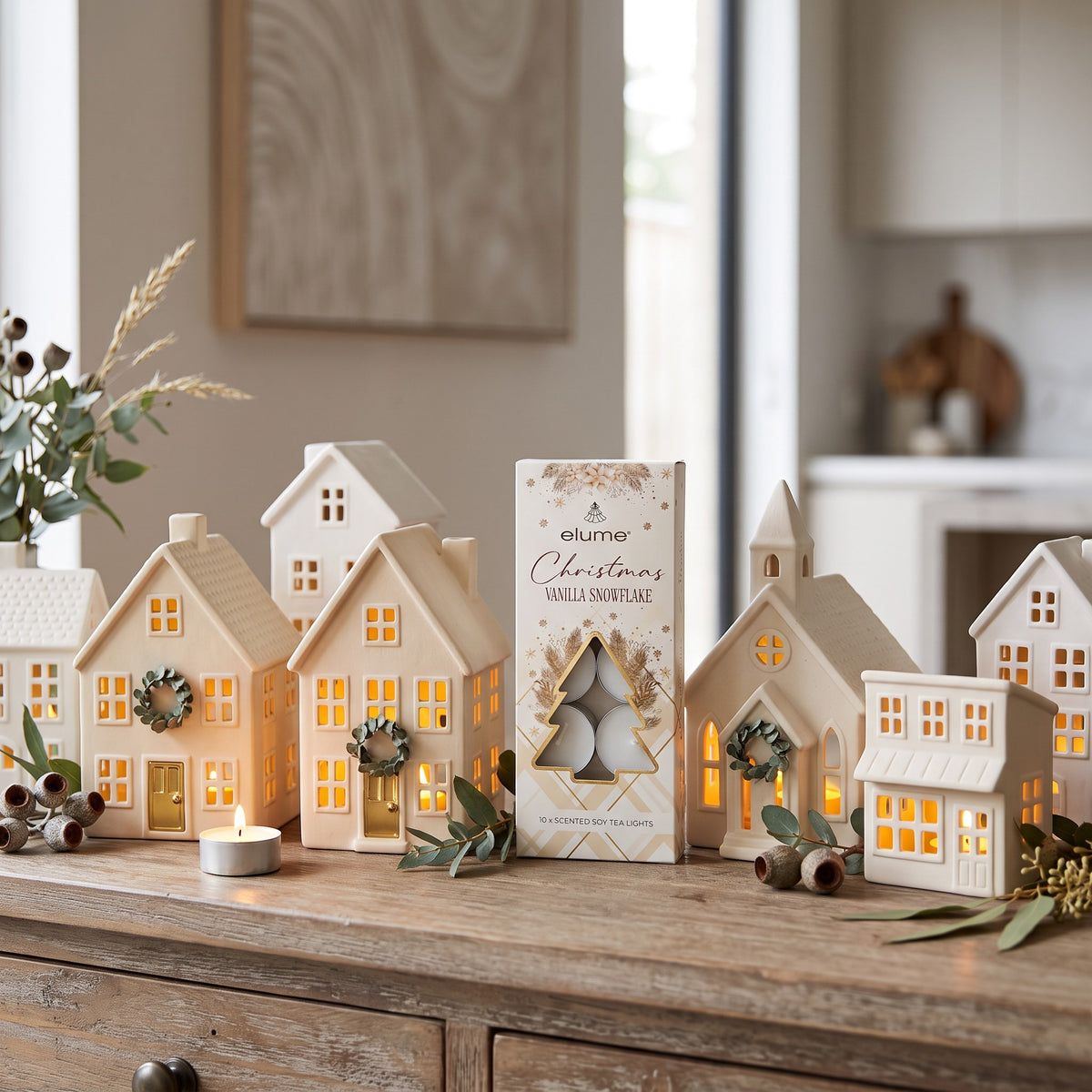 A set of Elume Christmas Vanilla Snowflake wax melts in a decorative house-shaped box, displayed on a rustic wooden table alongside lit tea light candles and greenery, with a softly blurred home interior background.